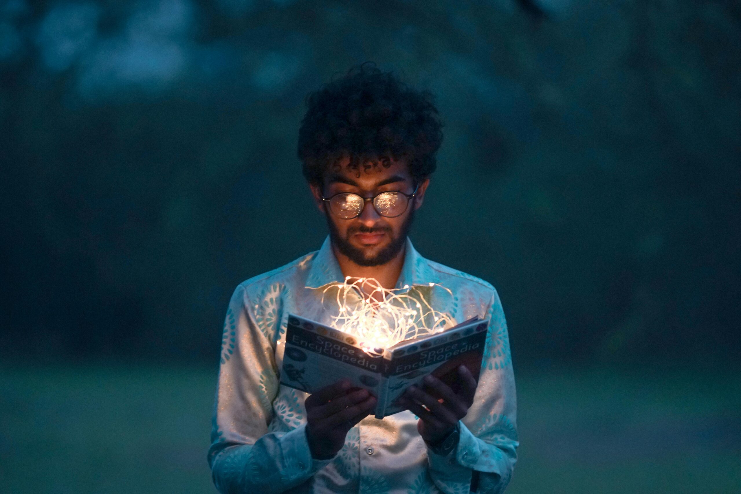 A man with glasses reads an illuminated book outdoors during twilight. Reading financial literacy